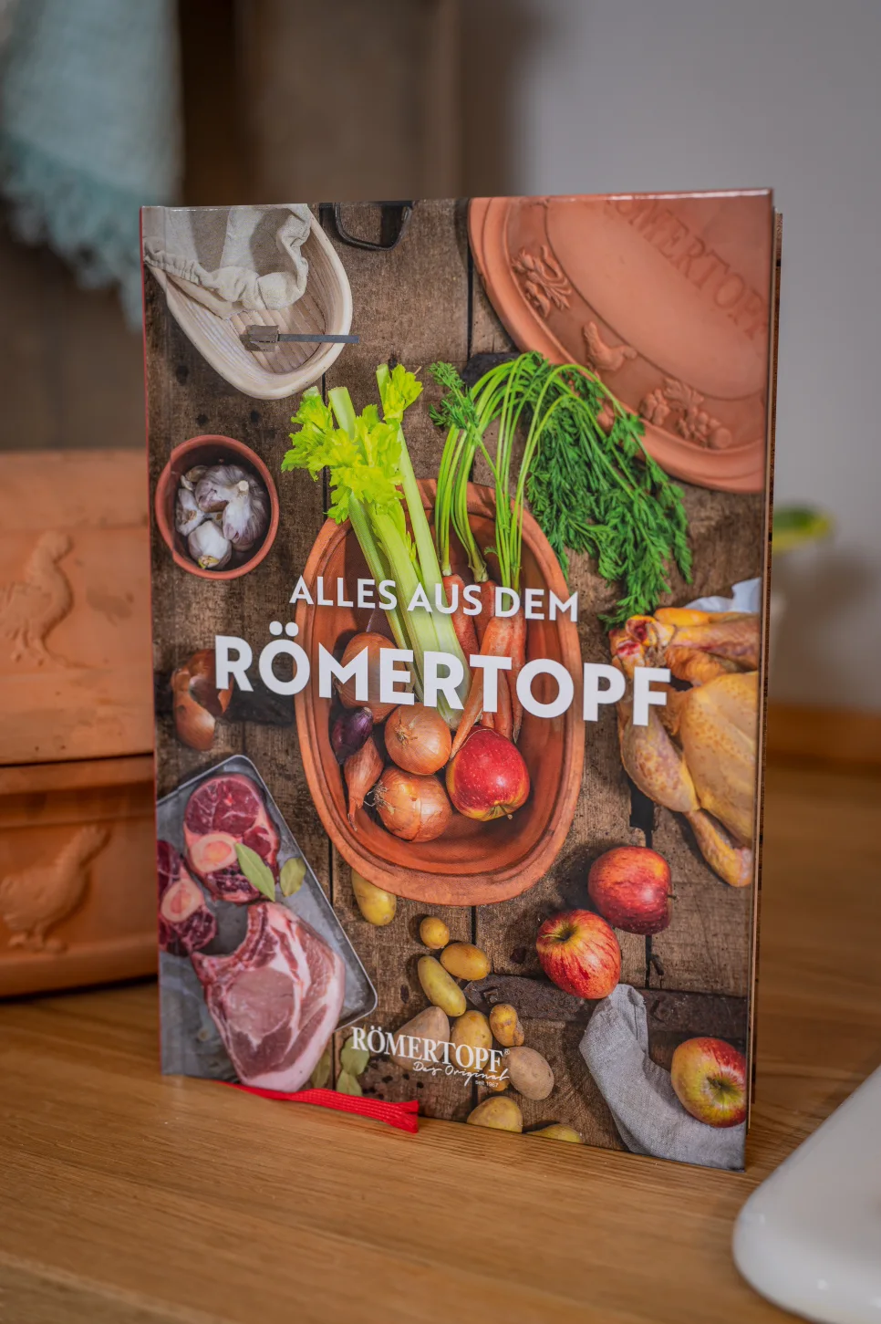 Römertopf Rezeptbuch – Inspiration und Rezepte für das Kochen im Tontopf Aufgeschlagenes Rezeptbuch mit Bildern und Texten, daneben ein unglasierter Römertopf sowie frische Zutaten wie Zitronen, Kräuter und Gewürze auf einem Holztisch arrangiert.