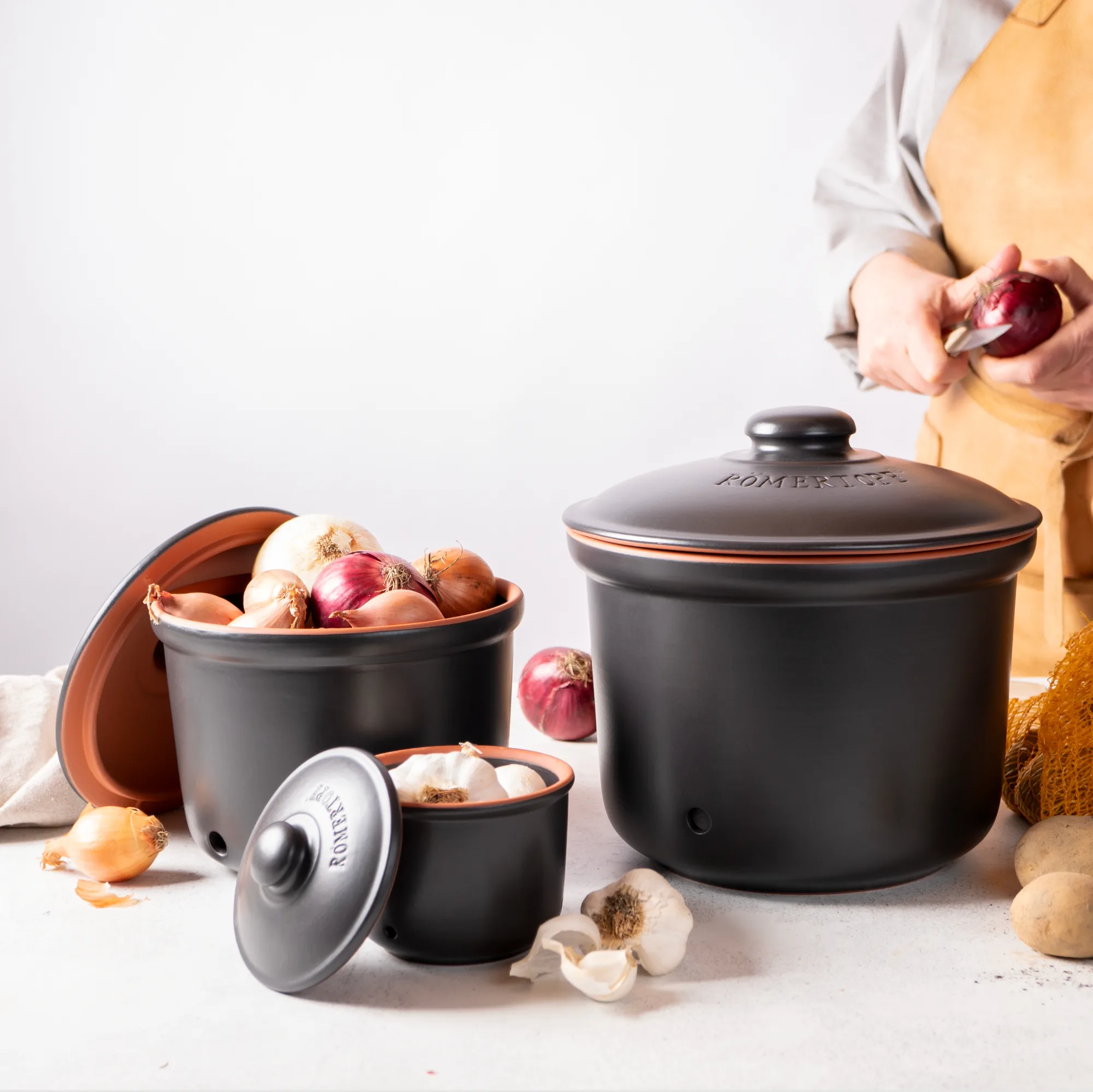 Römertopf Vorratstopf Set aus Keramik in Schwarz – 3-teiliges Aufbewahrungsgefäß für Küche Set aus zwei schwarzen Römertopf-Vorratstöpfen mit Deckel in verschiedenen Größen auf weißem Hintergrund; gestaltet zur atmungsaktiven Aufbewahrung von Lebensmitteln wie Zwiebeln oder Knoblauch.