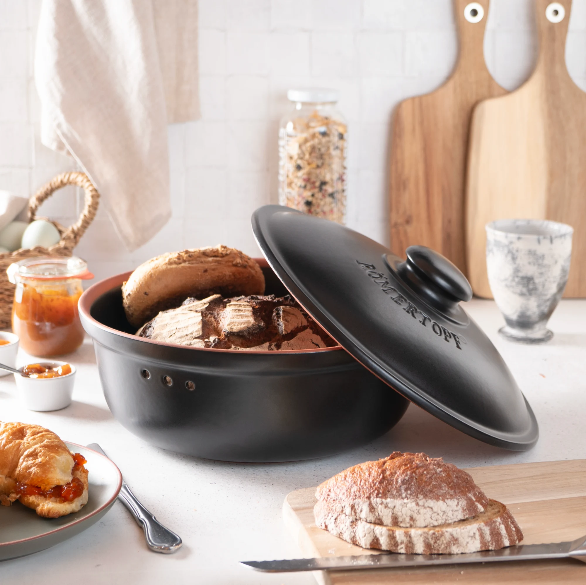 Römertopf Brottopf rund aus Keramik in Schwarz – Frischebehälter für Brot und Brötchen Runder, schwarzer Römertopf Brottopf mit Deckel aus atmungsaktiver Keramik, auf einem hellen Untergrund stehend. Der Deckel besitzt seitliche Griffrillen und Lüftungsöffnungen für optimale Luftzirkulation zur Brotaufbewahrung.