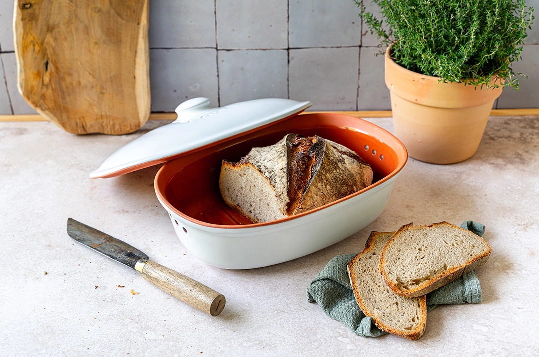 Römertopf Frische Töpfe aus Naturton zur nachhaltigen Aufbewahrung von Obst und Gemüse Drei Römertopf-Frischetöpfe aus Keramik in unterschiedlichen Größen und Farben (Weiß, Grün, Terrakotta) stehen nebeneinander auf einem Holzbrett; sie sind mit Deckeln verschlossen, darunter liegt Gemüse wie Zwiebeln und Knoblauch.