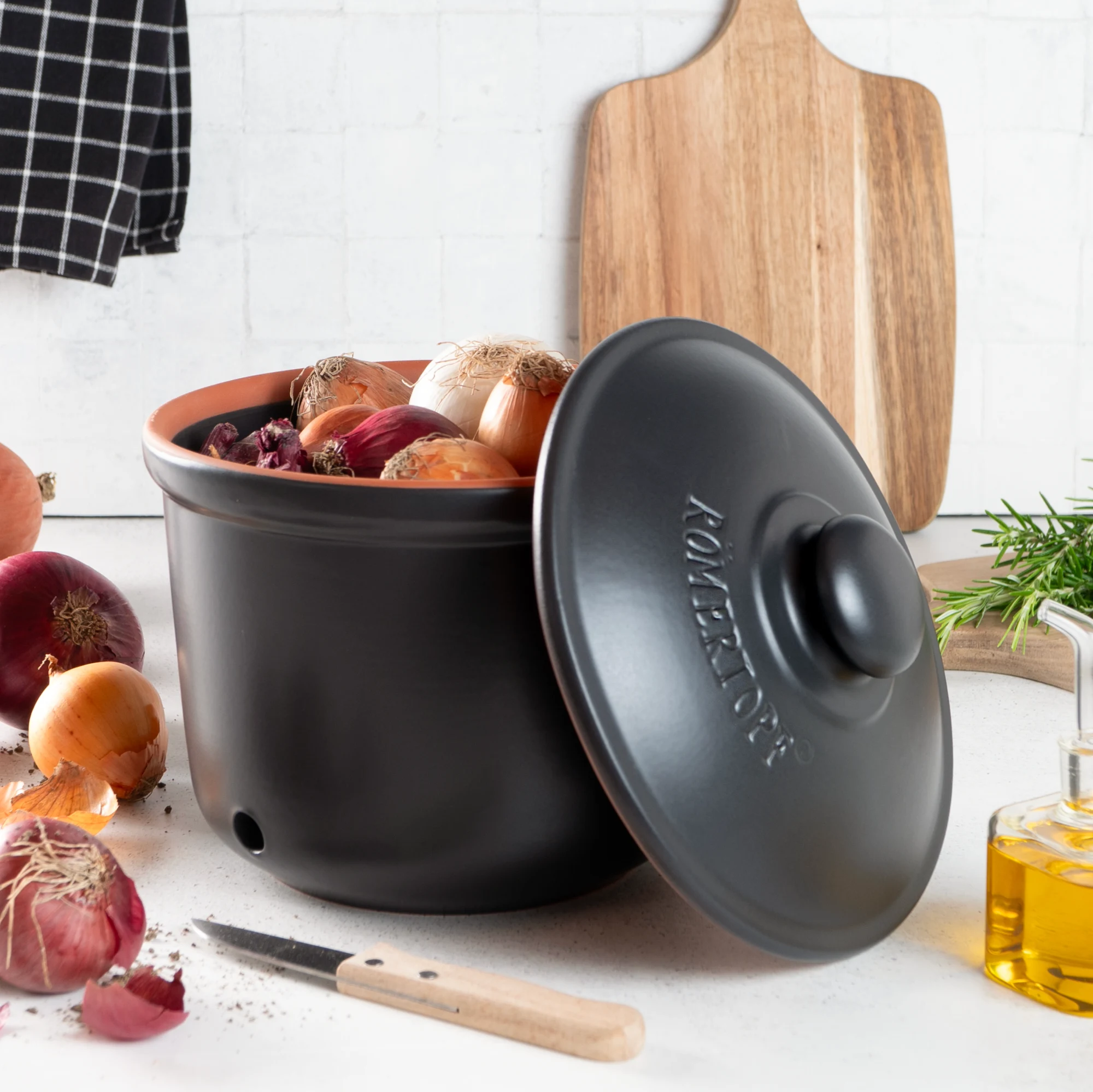 Römertopf Frischetopf Maxi Zwiebeltopf Schwarz – atmungsaktiver Keramiktopf zur Zwiebelaufbewahrung Schwarzer Römertopf-Zwiebeltopf in konischer Form mit mehreren seitlichen Luftlöchern, gefüllt mit braunen Zwiebeln; daneben liegen weitere Zwiebeln auf einer hellen Oberfläche vor neutralem Hintergrund.