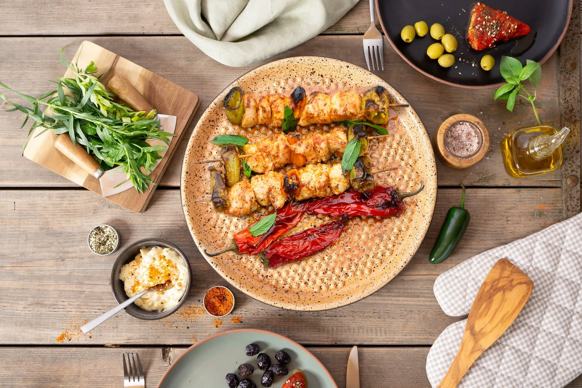 Grillplatte aus Keramik mit Gemüse und Fleisch auf dem BBQ Grill – Römertopf BBQ-Serie Ein Römertopf BBQ-Grill aus naturrotem Ton steht auf einem Holztisch im Freien, daneben liegen Gemüse, Kräuter und Grillzubehör; im Hintergrund sind Gartenmöbel und grüne Pflanzen sichtbar.