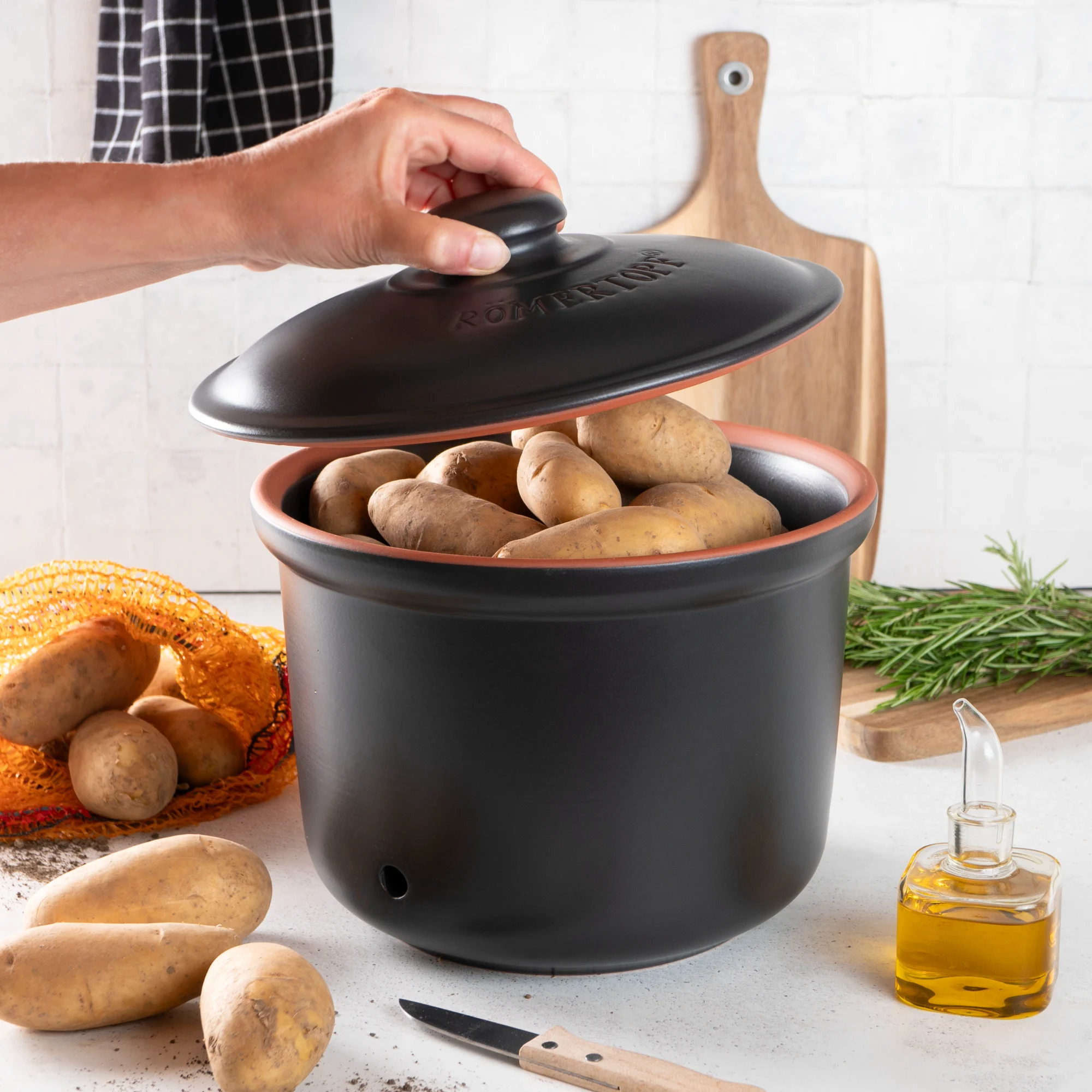Schwarzer Römertopf Kartoffeltopf aus Keramik mit Deckel – atmungsaktiv und ideal zur Lagerung von Kartoffeln Schwarzer Römertopf-Kartoffeltopf aus Keramik mit Deckel, stehend auf einer hellen Oberfläche; daneben liegen einige frische, ungekochte Kartoffeln.