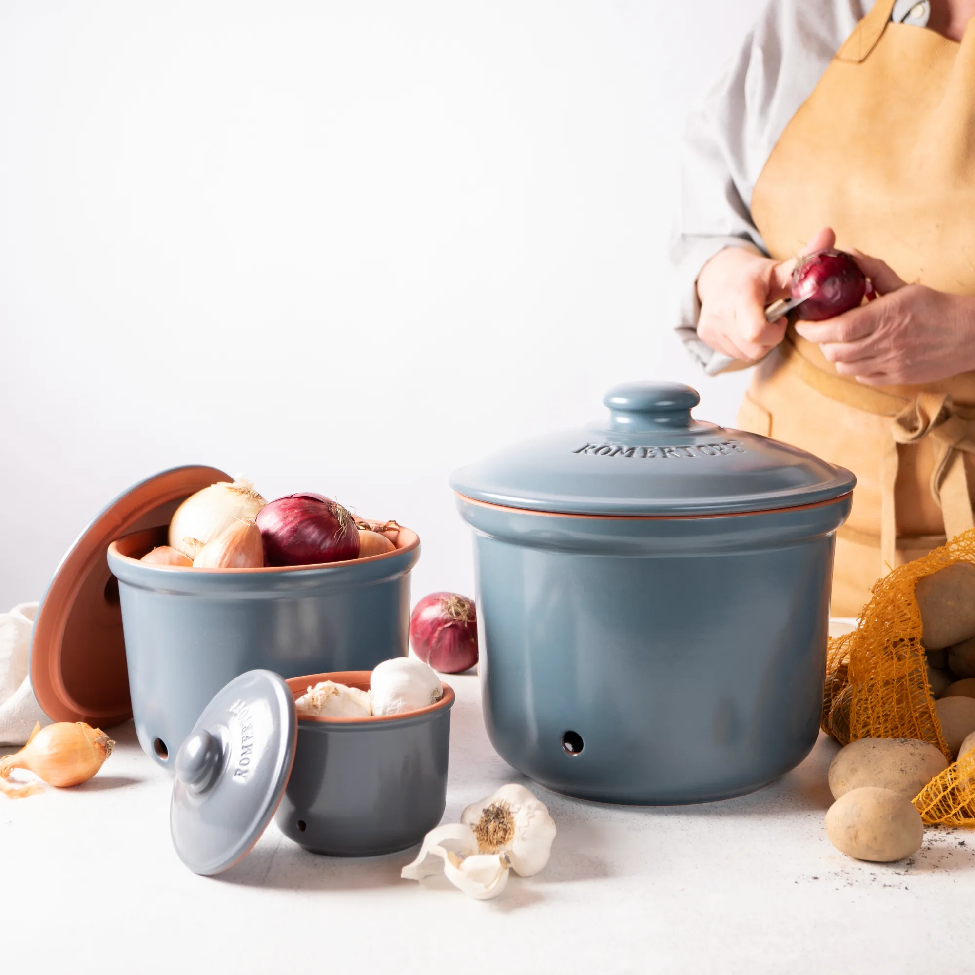 Römertopf Vorratstopf Set in Blaugrau – Keramik-Töpfe zur luftdichten Aufbewahrung von Lebensmitteln Set aus drei blaugrauen Römertopf-Vorratstöpfen aus Keramik mit Holzdeckeln, gestapelt auf einem Holztisch; jeder Topf ist mit einem weißen Schriftzug für „Knoblauch“, „Zwiebeln“ und „Kartoffeln“ gekennzeichnet.