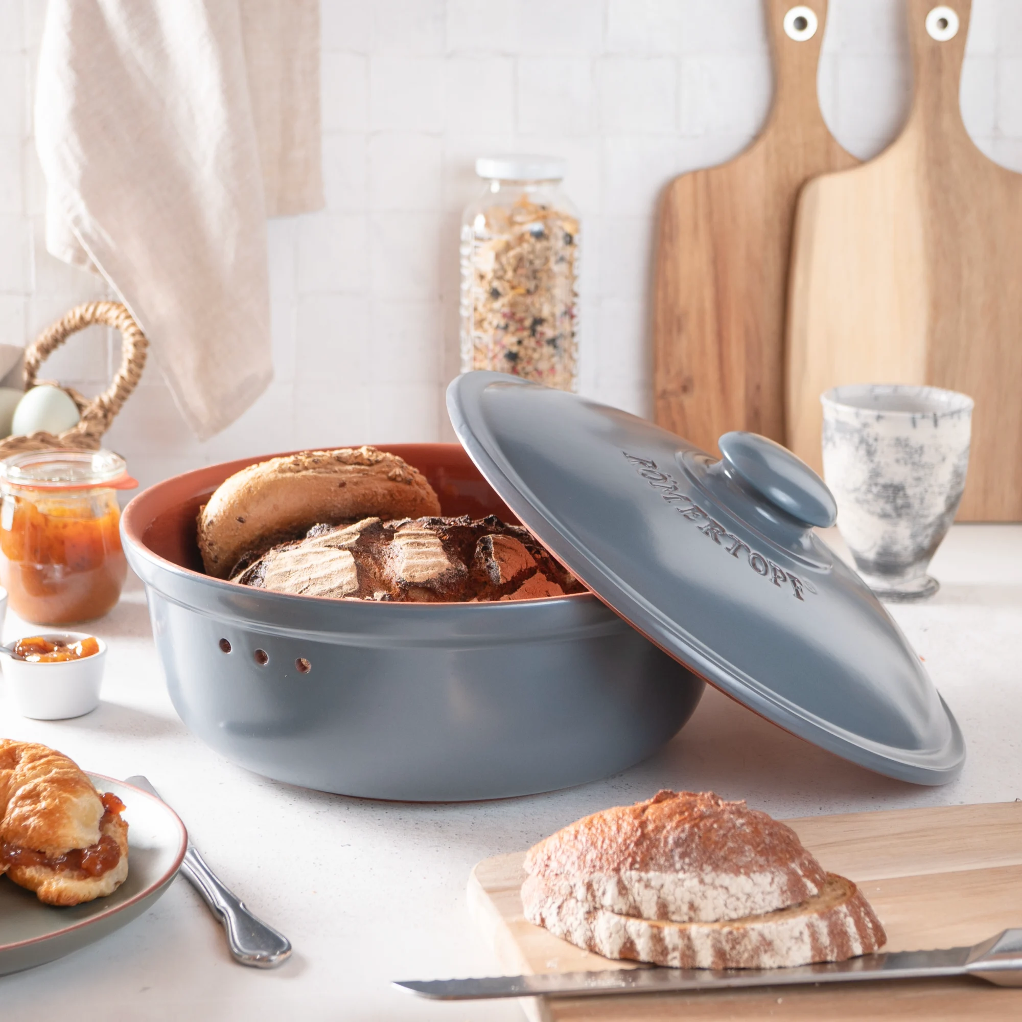 Römertopf Brottopf rund blaugrau – Frischetopf aus Naturton zur Brotaufbewahrung Runder Brottopf aus Keramik in Blaugrau mit passendem Deckel, Vorderseite mit Aufschrift „Römertopf“, stehend auf heller Oberfläche vor neutralem Hintergrund.