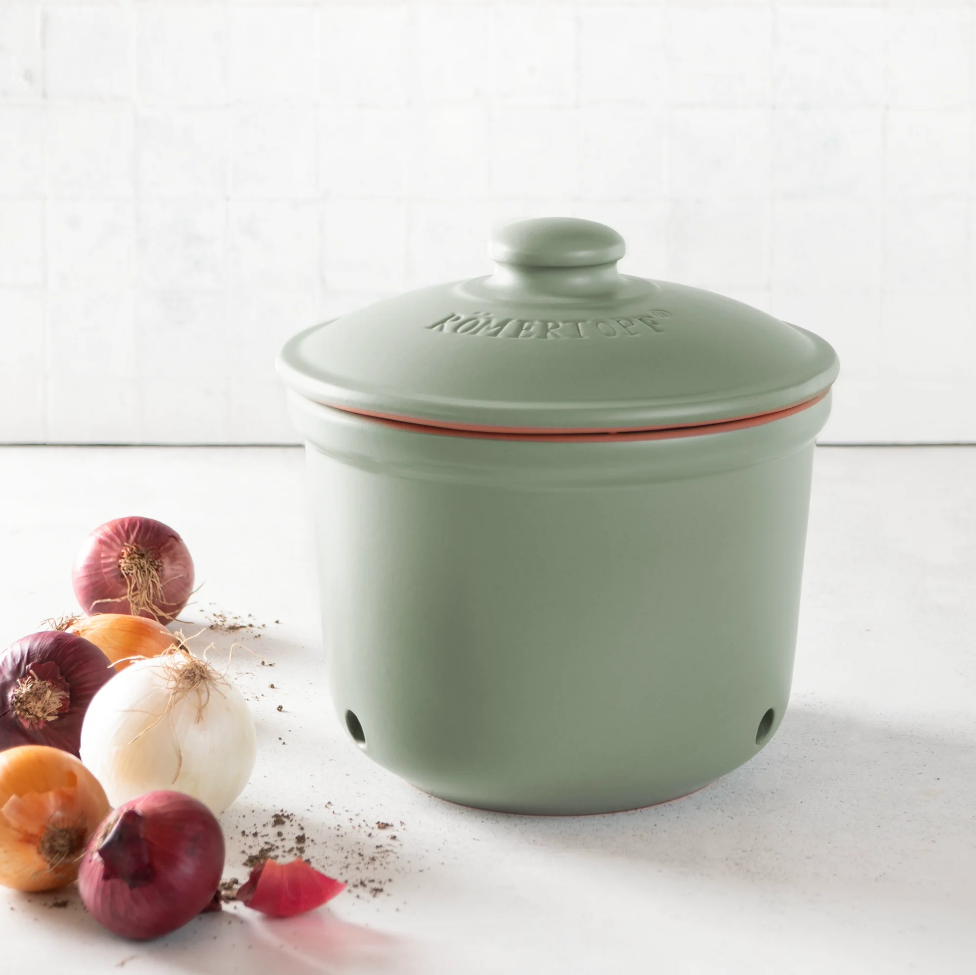 Römertopf Zwiebel-Frischetopf aus Keramik in Grün – luftdicht, atmungsaktiv, zur optimalen Lagerung Grüner Römertopf Zwiebel-Frischetopf aus Keramik mit Deckel und Belüftungslöchern, auf einem hellen Untergrund stehend.