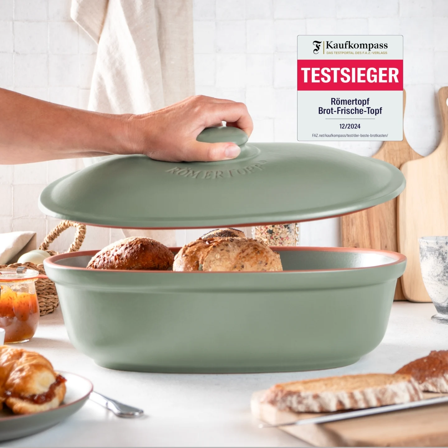 Frischetopf Maxi Grün für Brotaufbewahrung – Römertopf Vorratsdose aus Keramik Grüner Römertopf Brot-Frischetopf Maxi mit gelochtem Deckel, stehend auf einem Holzuntergrund, im Hintergrund unscharfe Küchenutensilien.