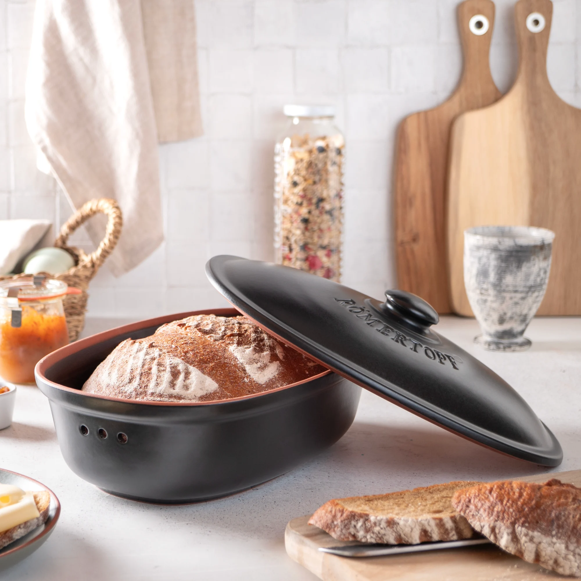 Römertopf Brottopf Frische Schwarz – Keramik Brotkasten für länger frisches Brot Schwarzer Römertopf-Frische-Brottopf mit Deckel aus Naturton, seitlich mit Lüftungslöchern und eingraviertem „Römertopf“-Schriftzug, vor weißem Hintergrund.