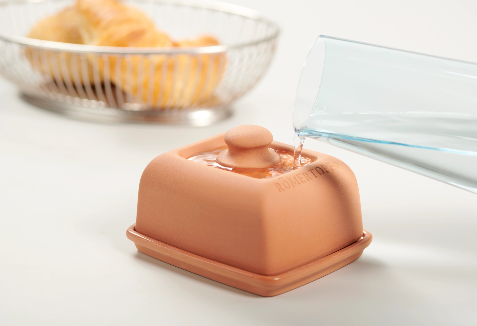 Römertopf Butterdose aus Keramik mit Klimafunktion – Frische Butter stilvoll aufbewahren Keramik-Butterdose von Römertopf in Naturton mit Deckel, auf einem hellen Untergrund; daneben liegt ein Holzlöffel, im Hintergrund ein Holzbrett mit Brot.