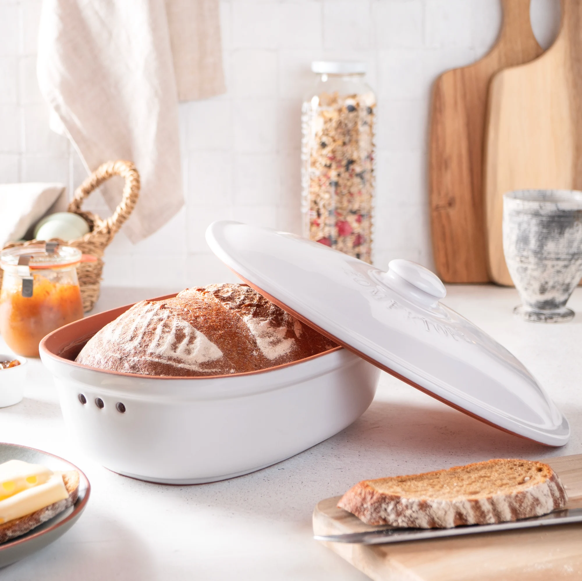 Römertopf Brottopf Frischetopf Medi aus Keramik in Weiß – Brot richtig aufbewahren Weiß glasierter Römertopf Brottopf mit Deckel auf einem Holztisch, im Inneren liegt ein rundes Brot; im Hintergrund steht ein weiterer Brottopfdeckel und etwas Küchenzubehör.