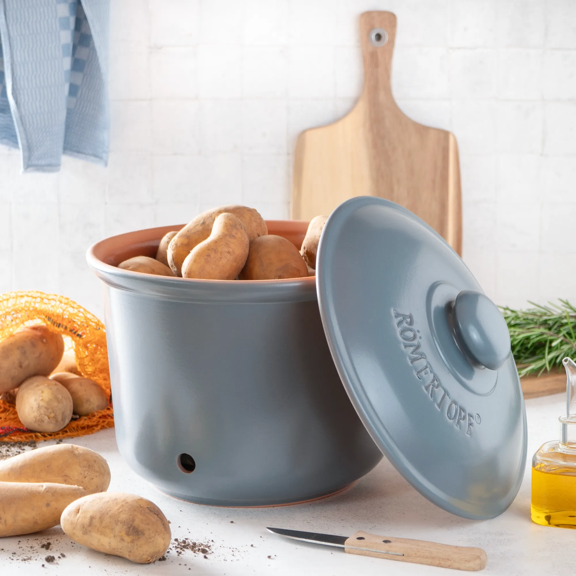 Römertopf Vorratstopf Set blaugrau aus Keramik – luftdicht & dekorativ zur Aufbewahrung von Lebensmitteln Zwei blaugraue Römertopf-Vorratstöpfe aus Keramik mit Deckeln stehen nebeneinander auf einer Küchenarbeitsplatte; die Töpfe sind unterschiedlich groß und tragen weiße Aufschriften „ZWIEBELN“ und „KNOBLAUCH“, Umgebung mit frischen Lebensmitteln dekoriert.