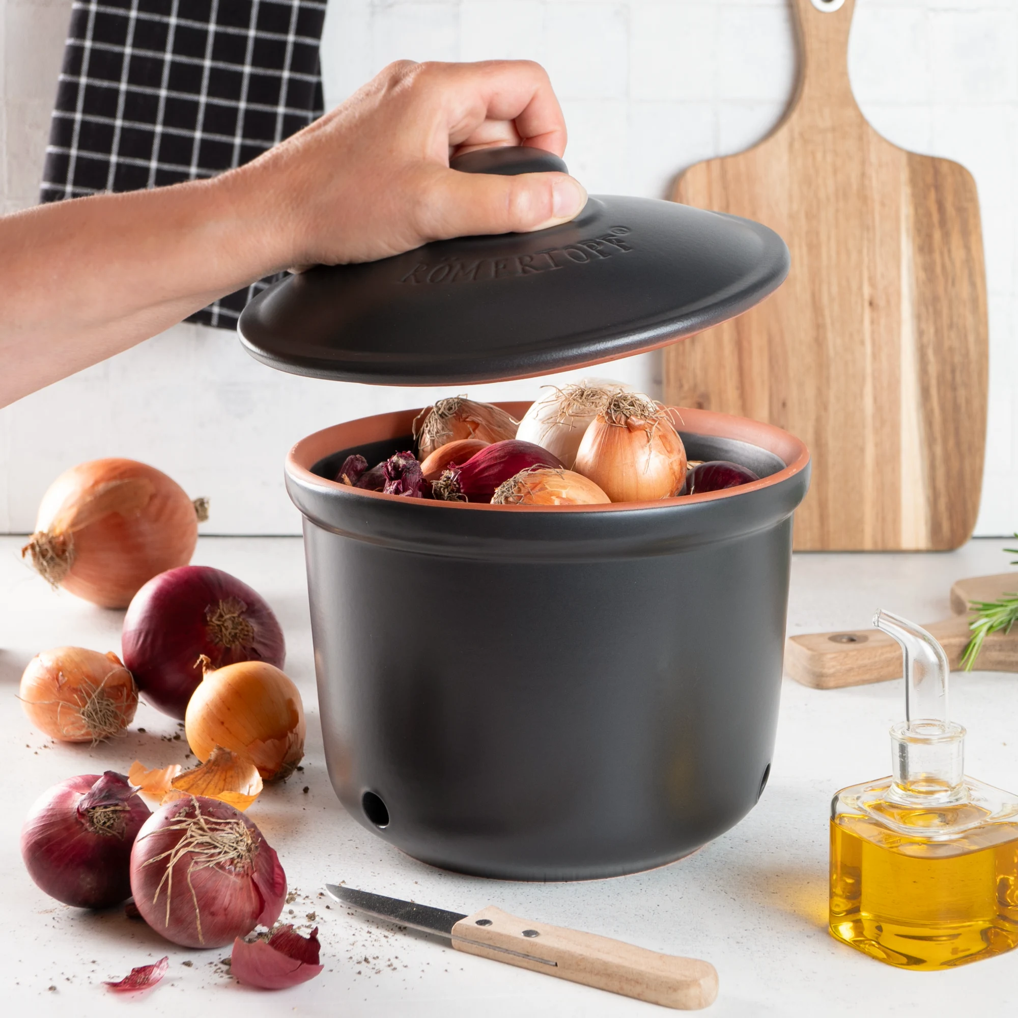 Römertopf Zwiebeltopf Maxi Schwarz – Frischetopf zur optimalen Lagerung von Zwiebeln Schwarzer Römer Topf Frischetopf Maxi aus Keramik mit Deckel, belüftet durch seitliche Luftlöcher, gefüllt mit roten Zwiebeln, stehend auf einer Arbeitsfläche.