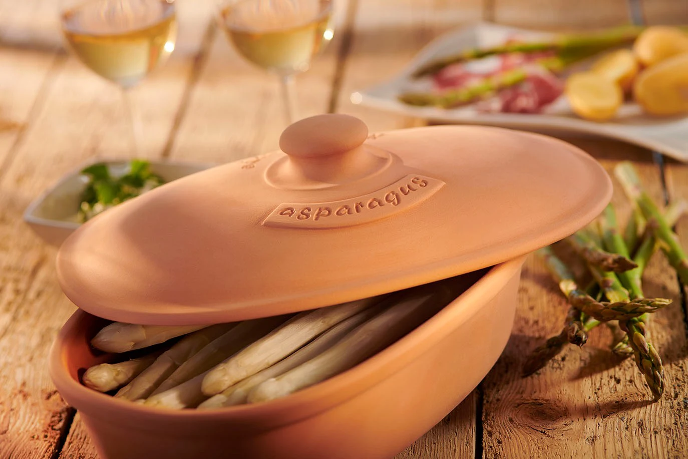 Spargel im Römertopf: Schonend gegartes Frühlingsgericht Ein Römertopf mit gegartem Spargel, Möhren und Zitronenscheiben, serviert auf einem hellen Untergrund mit Kräuterdekoration.