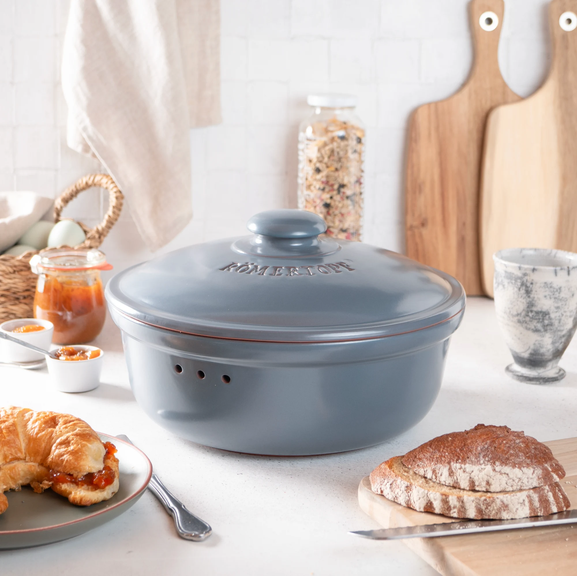 Römertopf Brottopf rund blaugrau – Frischebehälter aus Naturton für lange Brotfrische Runder Brottopf aus Keramik in Blaugrau mit passendem Deckel, seitlicher Luftzirkulation und Innengravur „Römertopf“, auf hellem Untergrund platziert.