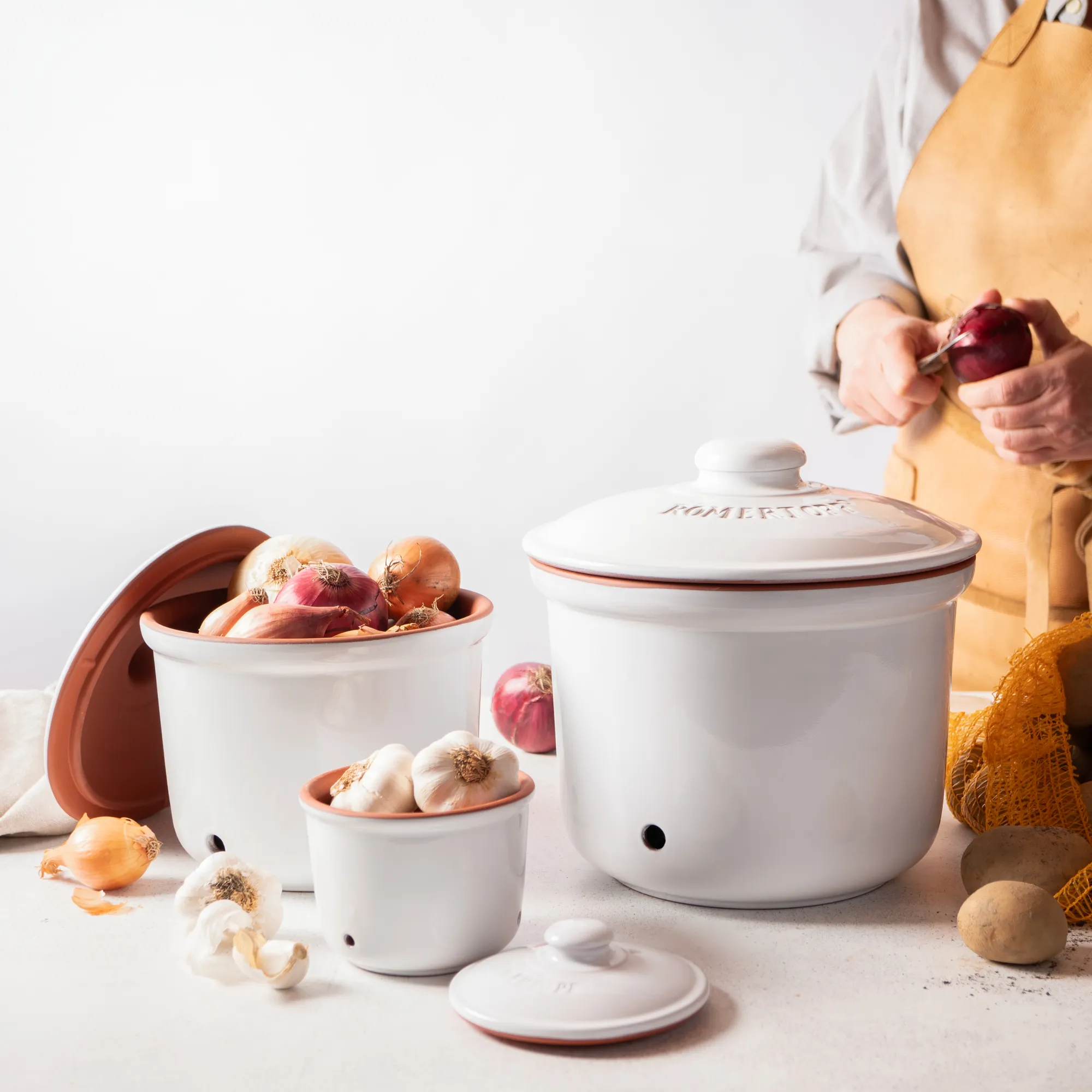 Römertopf Vorratstopf Set aus Keramik in Weiß – Luftdichtes Aufbewahren für Küche und Haushalt Vorratstopf-Set aus weißer Keramik von Römertopf, bestehend aus drei Töpfen mit Deckel in verschiedenen Größen, jeweils mit Beschriftung „Zwiebeln“, „Knoblauch“ und „Kartoffeln“, auf heller Oberfläche arrangiert.