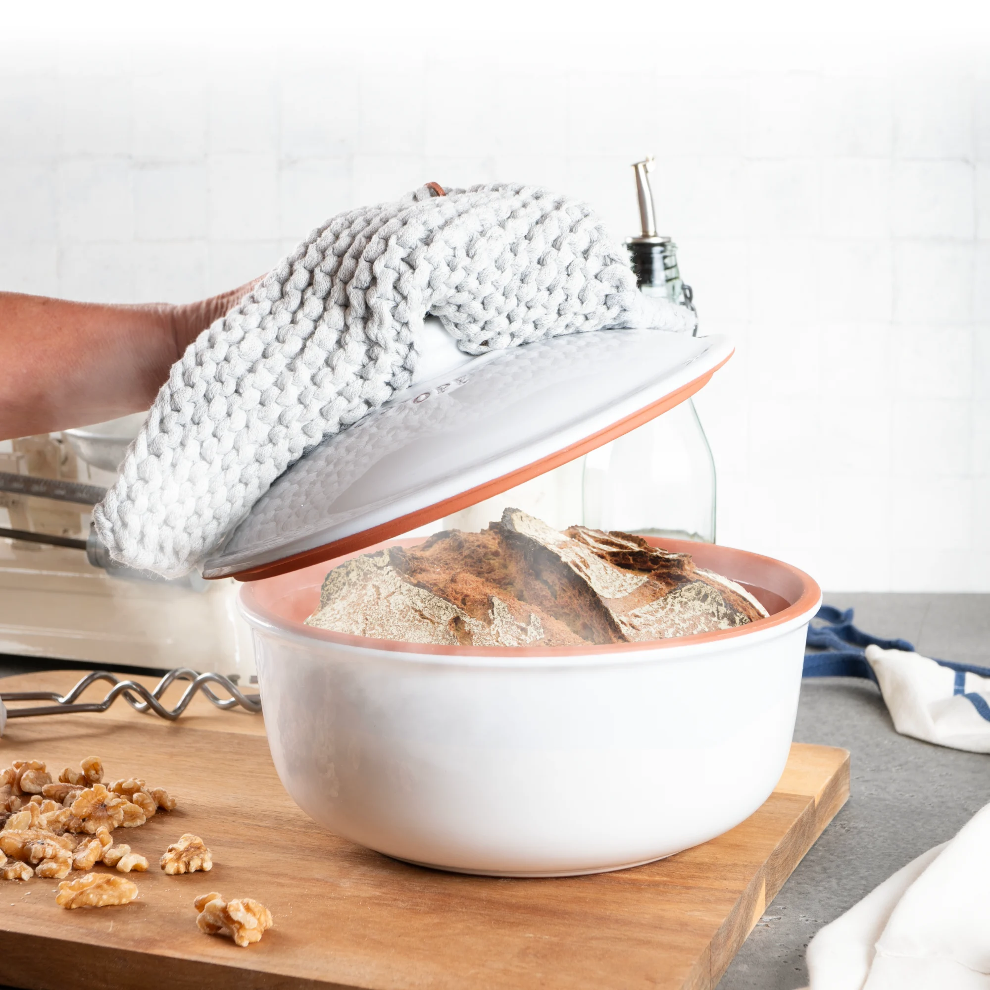 Römertopf Brotbacktopf aus Keramik in Weiß – ovaler Brottopf mit Deckel für traditionelle Brotherstellung Weißer Römertopf Brot-Backtopf mit Deckel auf einem Holztisch, im Hintergrund ein gebackenes Brot und ein Messer.