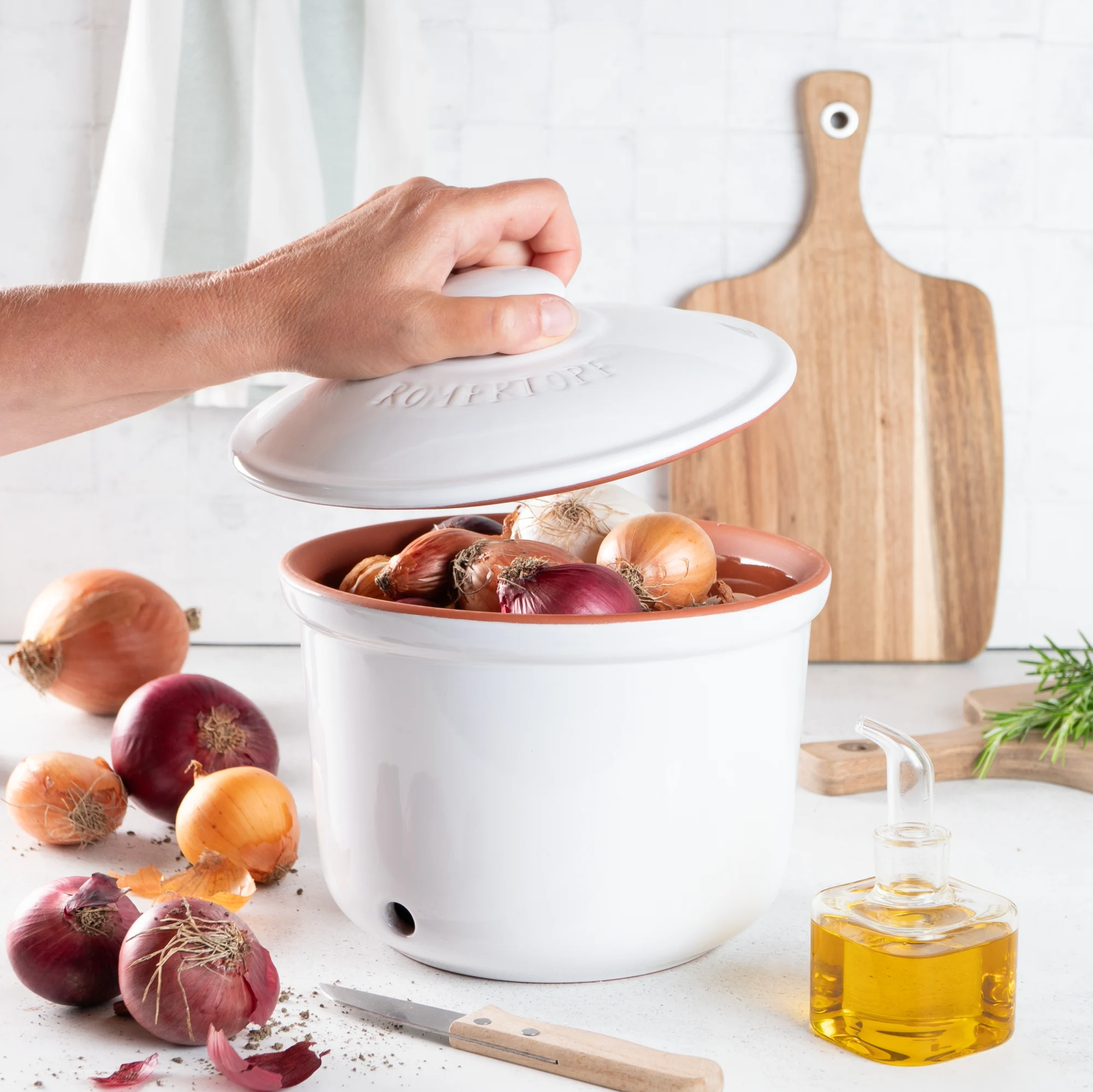 RÖMERTOPF Zwiebeltopf Maxi Weiß – Keramik Frischetopf zur Zwiebel-Aufbewahrung Weißer Römertopf-Zwiebeltopf aus Keramik mit Deckel, belüftet durch kleine Löcher, steht auf einer Küchenarbeitsplatte; daneben liegen zwei Zwiebeln.