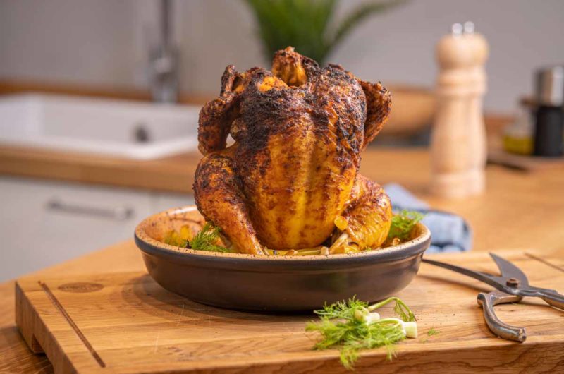 Gebackenes Hähnchen in einer runden schwarzen Pfanne, präsentiert auf einem Holztisch in einer Küche.