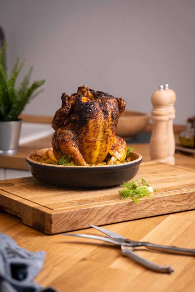 Gebackenes Hähnchen aufrecht in einer Schale auf Holzbrett, im Hintergrund Pfeffermühle und Pflanze.