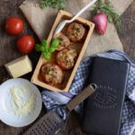 Gefüllte Tomaten in einer Keramikform, umgeben von Kräutern, Tomaten, Zwiebeln, Käse und einer Reibe auf Holzuntergrund.