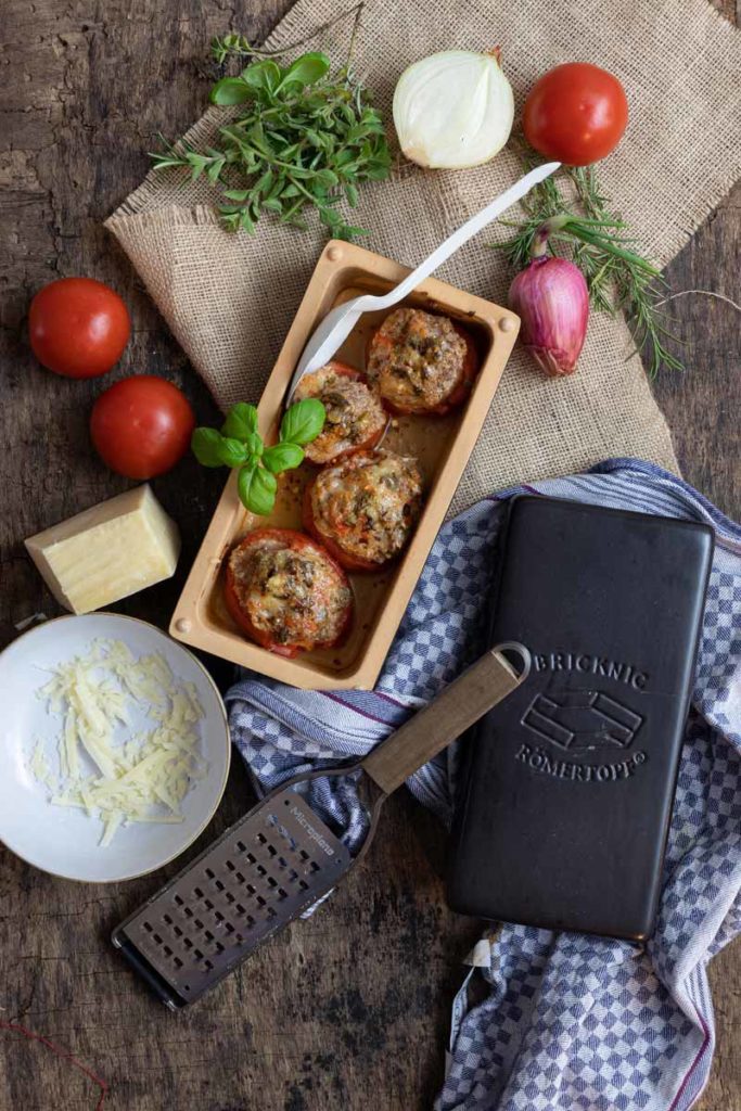 Gefüllte Tomaten in einer Keramikform, umgeben von Kräutern, Tomaten, Zwiebeln, Käse und einer Reibe auf Holzuntergrund.