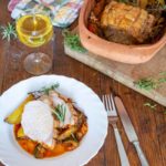 Weißer Teller mit Braten, Ofengemüse und Rosmarin, daneben ein Glas Weißwein, Gabel und Messer auf Holztisch.
