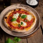 Pizza Margherita mit frischem Basilikum auf einem Teller, daneben Olivenöl und eine Schale Mehl auf Holztisch.