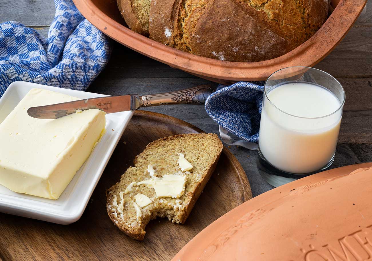 Brot mit Butter, Messer, ein Glas Milch und eine Brotscheibe auf Holzbrett. Blauer Stoff und Brottopf im Hintergrund.