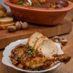 Teller mit Hähnchenkeule in Gemüse-Soße und Baguettescheiben, im Hintergrund ein gefüllter Bräter.
