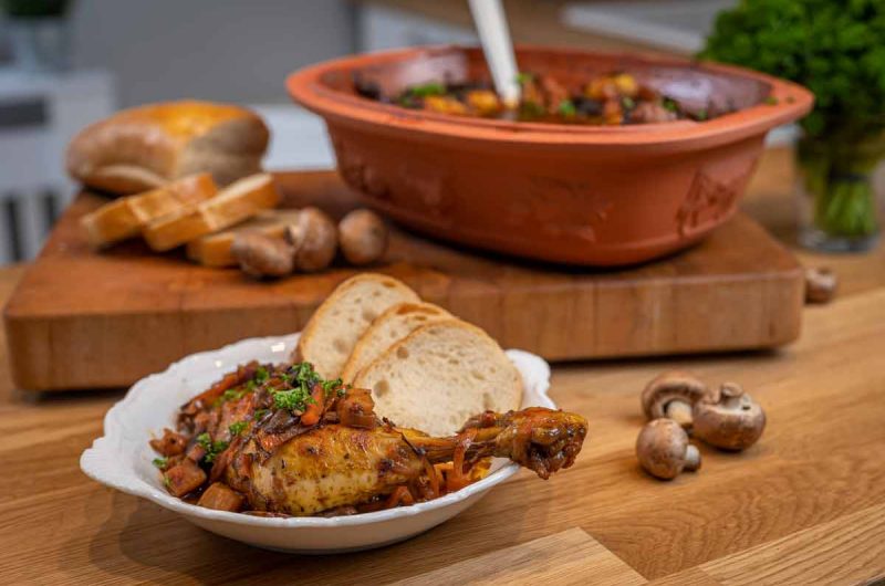 Hähnchenschenkel in Sauce mit Brot und Pilzen auf einem Teller. Tontopf im Hintergrund auf Holzbrett.