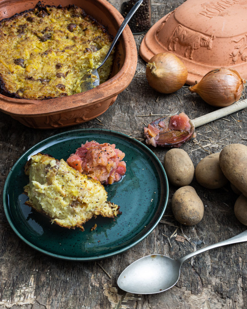 Ton-Schmortopf mit Kartoffelauflauf, serviert auf Teller mit Apfelmus; daneben Zwiebeln, Kartoffeln und ein Löffel.