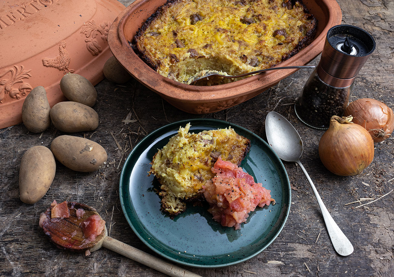 Kartoffelauflauf mit Zwiebeln und Apfelkompott auf Teller, umgeben von Kartoffeln und Gewürzen auf Holzuntergrund.
