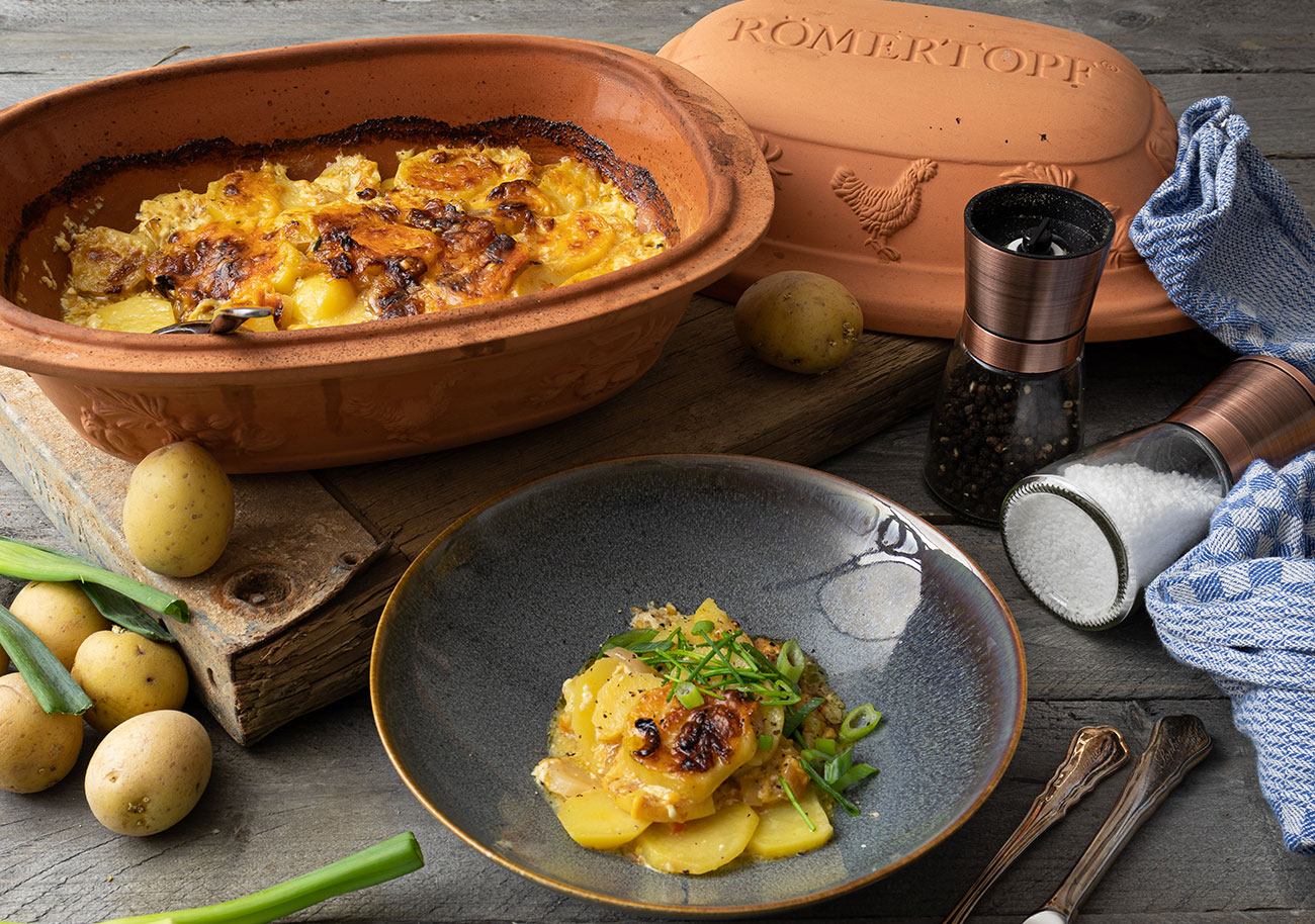 Kartoffelgratin im Römertopf, serviert mit Frühlingszwiebeln. Zutaten: Kartoffeln, Pfeffer, Salz. Teller und Gewürze daneben.