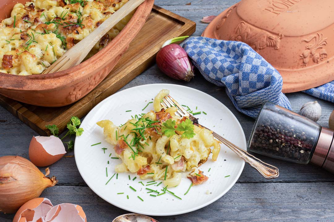 Auflauf mit Nudeln und gebräuntem Käse auf Teller und in Römertopf, umgeben von Zwiebeln, Gewürzen und Geschirrtuch.