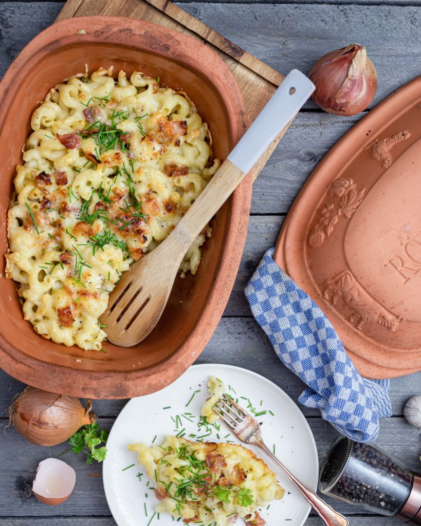 Auflaufform mit überbackenen Makkaroni, Schinken und Kräutern; Servierlöffel aus Holz und gefüllter Teller auf Holztisch.