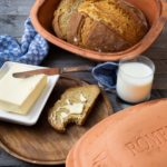 Brot im Tontopf, Brot mit Butter, Messer, Milchglas und blaues Handtuch auf Holztisch.