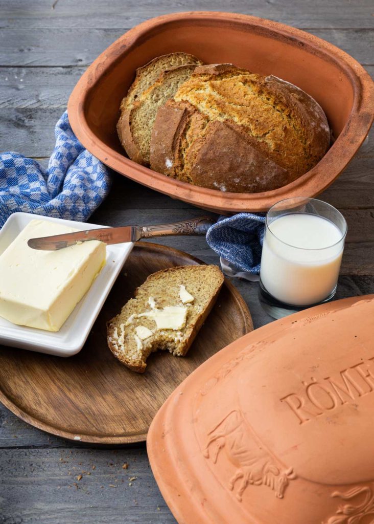 Brot im Tontopf, Brot mit Butter, Messer, Milchglas und blaues Handtuch auf Holztisch.