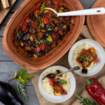 Keramikschale mit Ratatouille, angerichtet mit Couscous in Schalen, daneben frisches Gemüse und Kräuter auf Holztisch.