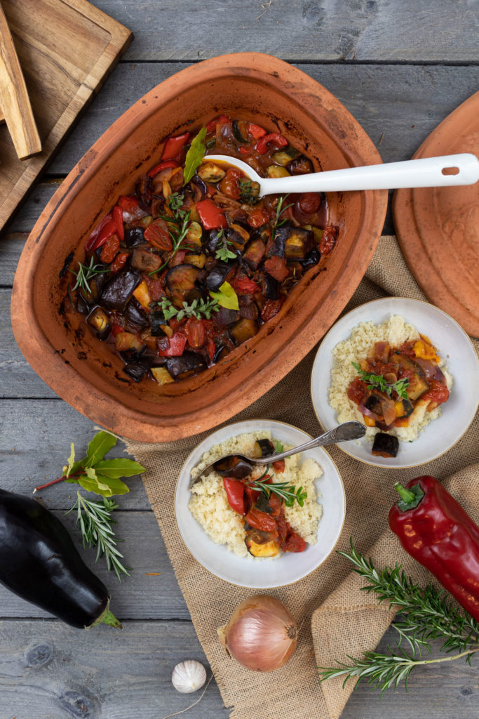 Keramikschale mit Ratatouille, angerichtet mit Couscous in Schalen, daneben frisches Gemüse und Kräuter auf Holztisch.