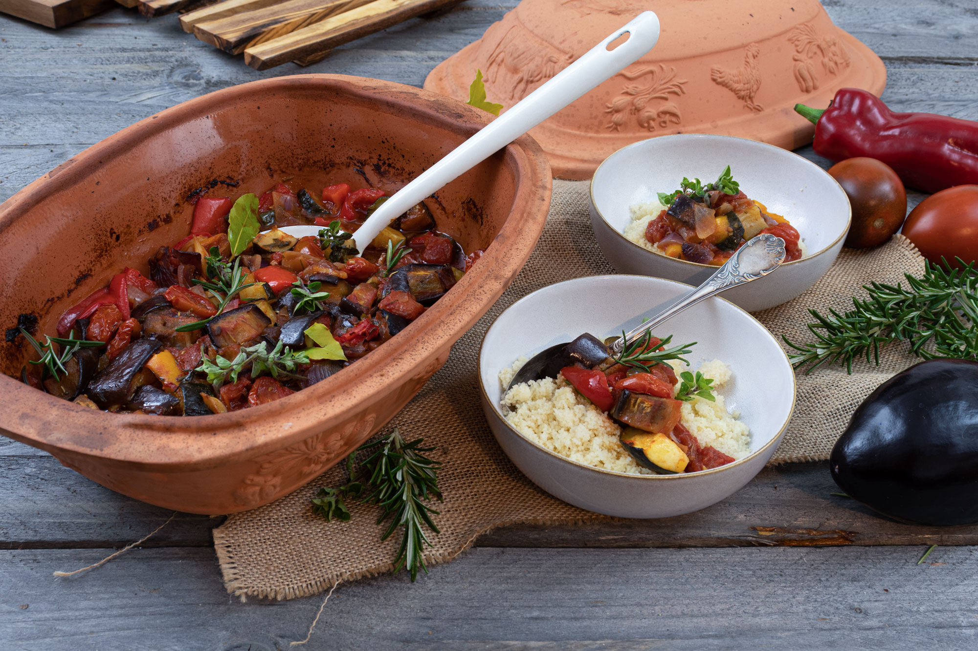 Tonpfanne mit Ratatouille, zwei Schalen mit Couscous und Gemüse, dekoriert mit frischen Kräutern auf Holzuntergrund.