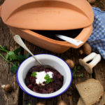 Ein Römertopf auf rustikalem Holz, daneben ein Teller Gulasch mit Sauerrahm und Kräutern, Brot und Pilze.