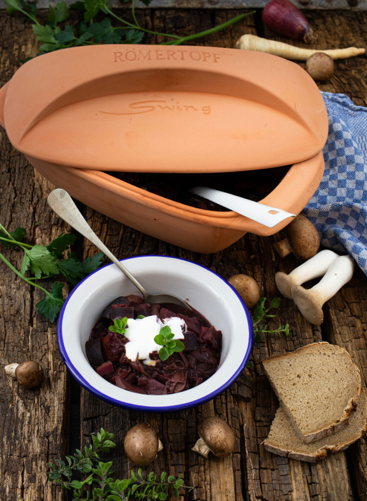 Ein Römertopf auf rustikalem Holz, daneben ein Teller Gulasch mit Sauerrahm und Kräutern, Brot und Pilze.