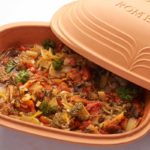 Ton-Römertopf mit Gemüseeintopf; Brokkoli, Paprika, Tomaten und Kräuter sichtbar, Topfdeckel leicht geöffnet.