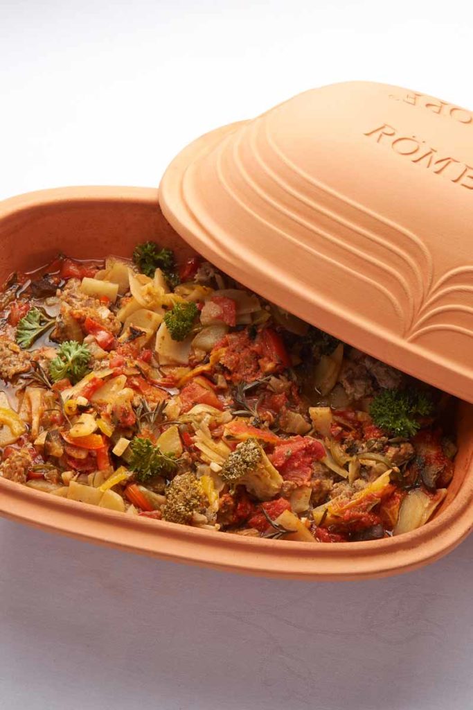 Ton-Römertopf mit Gemüseeintopf; Brokkoli, Paprika, Tomaten und Kräuter sichtbar, Topfdeckel leicht geöffnet.