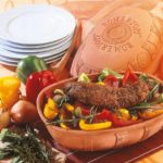 Römertopf mit Fleisch und buntem Gemüse, dekoriert mit Kräutern, daneben Paprika, Zwiebeln und Geschirrstapel.