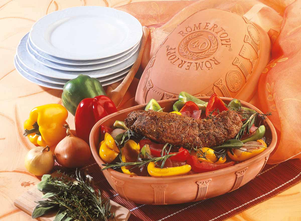 Römertopf mit Fleisch und buntem Gemüse, dekoriert mit Kräutern, daneben Paprika, Zwiebeln und Geschirrstapel.