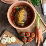 Auf einem Holztisch steht ein Tontopf mit Hackbraten und Tomaten. Daneben liegen Gemüse, ein Messer und Holzkochlöffel.