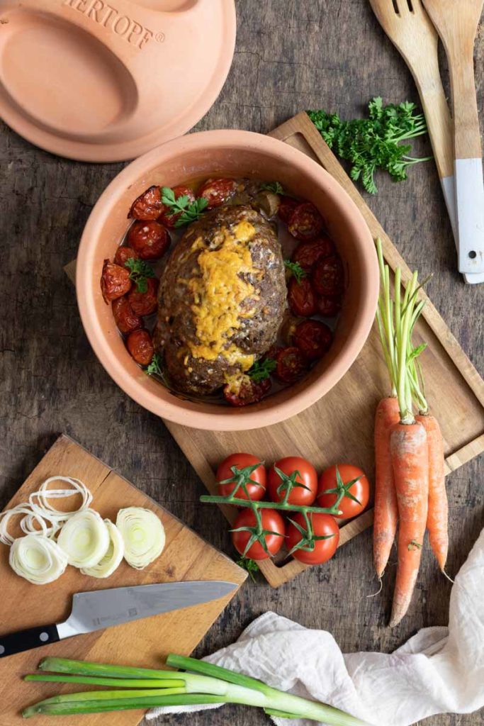 Auf einem Holztisch steht ein Tontopf mit Hackbraten und Tomaten. Daneben liegen Gemüse, ein Messer und Holzkochlöffel.