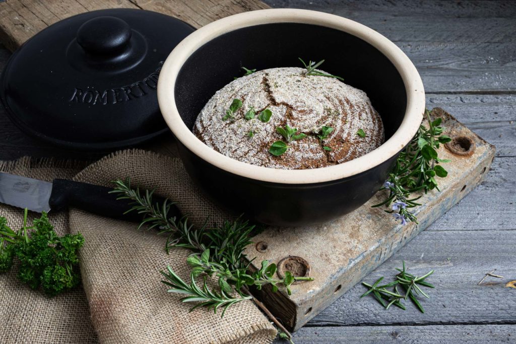 Frisch gebackenes Brot in einem Römertopf auf Holzbrett, umgeben von Kräutern und einem Messer.