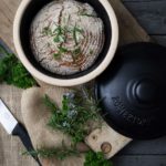 Rundes Brot in einem Römertopf, dekoriert mit frischen Kräutern, auf einem Holzbrett und Jutesack neben einem Messer.